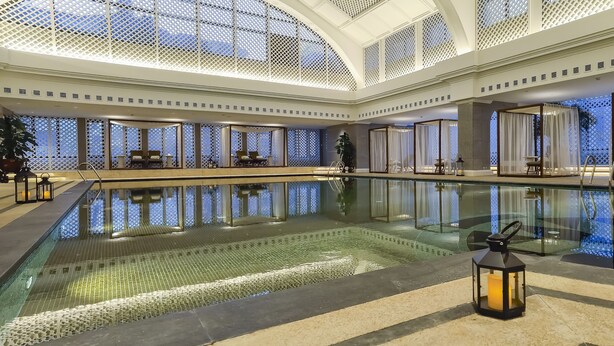 Una amplia piscina interior con agua cristalina, rodeada de zonas de descanso y un techo alto con celosías decorativas.