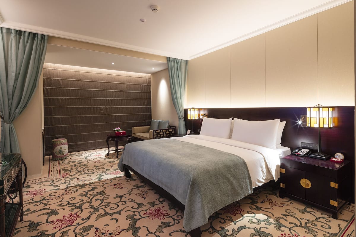 A hotel room with a large bed, a nightstand, a lamp, a small table, and a decorative rug.