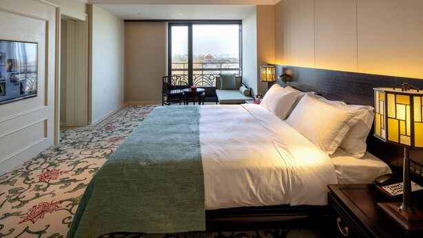 A hotel room with a large bed, a desk with a chair, a window with a view, and a nightstand with a lamp. - Grand Hotel Beijing   (Beijing)