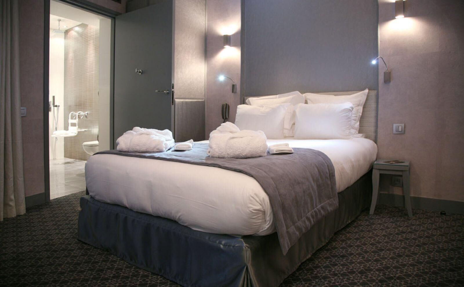 A hotel room with a bed, a bedside table, a bathroom visible through an open door, and wall-mounted lighting.