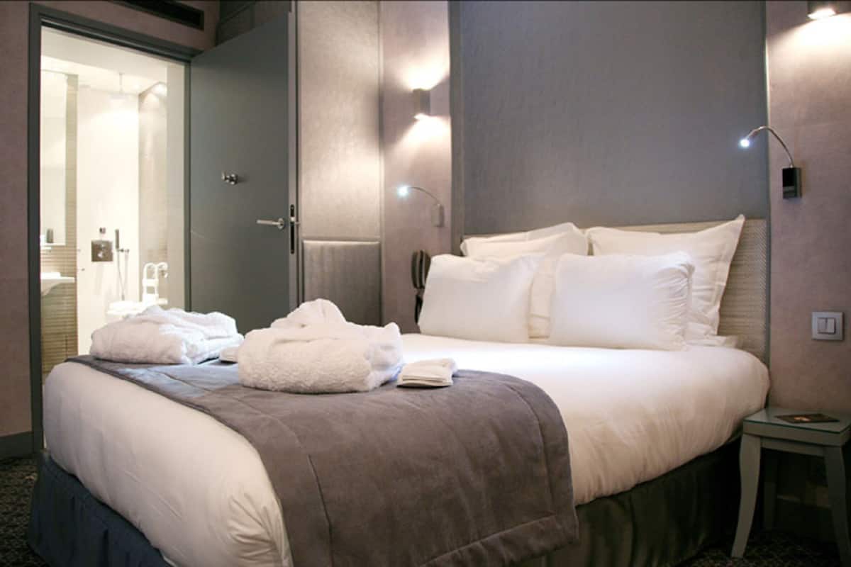 Une chambre d’hôtel avec un grand lit, une table de chevet, une salle de bain visible à travers une porte ouverte et un luminaire fixé au mur.