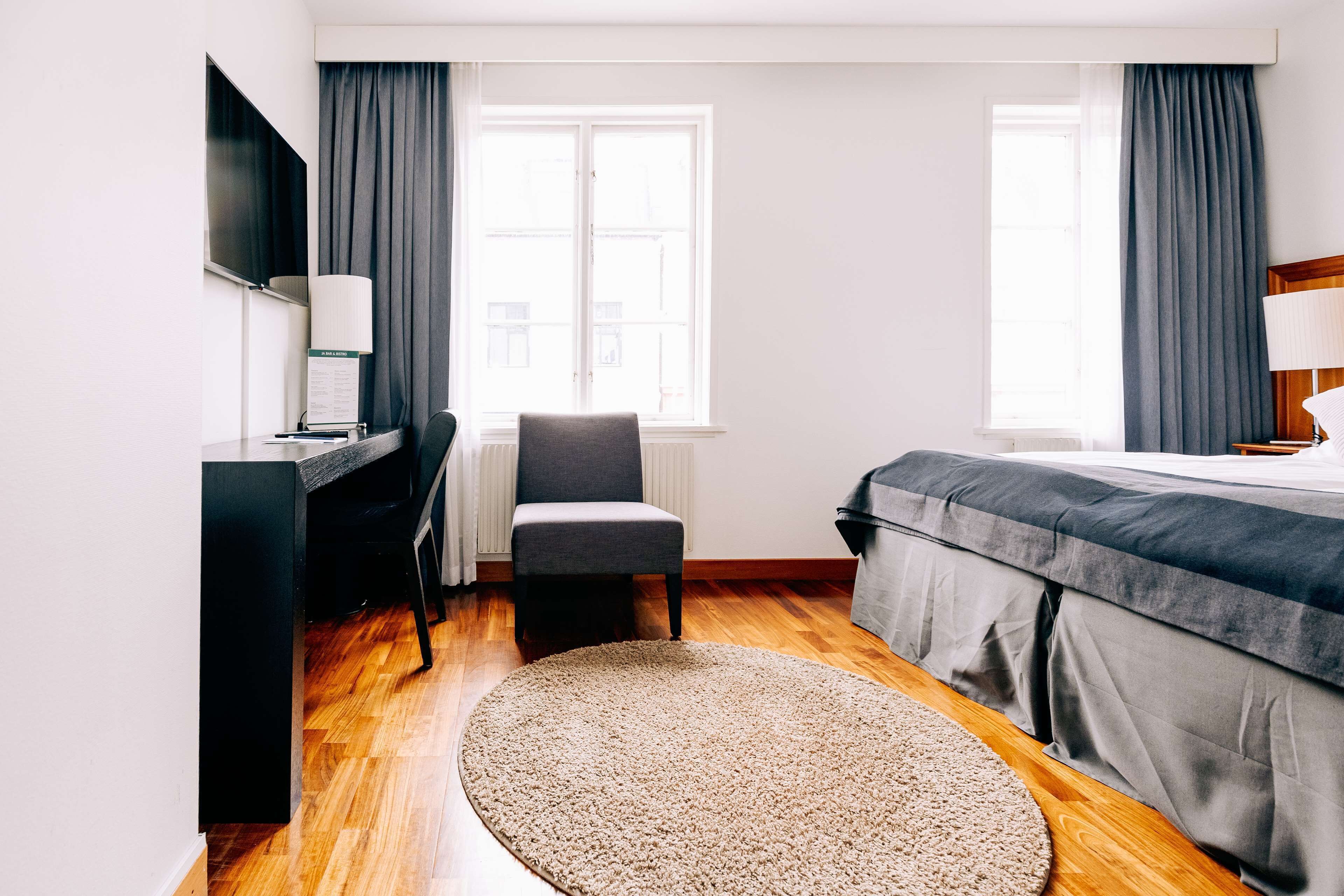 Ein Hotelzimmer mit Schreibtisch, Stuhl, Bett und zwei Fenstern mit Vorhängen.