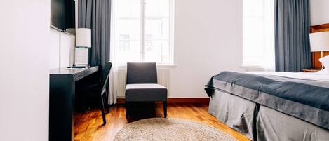 A hotel room with a desk, chair, bed, and two windows with curtains.