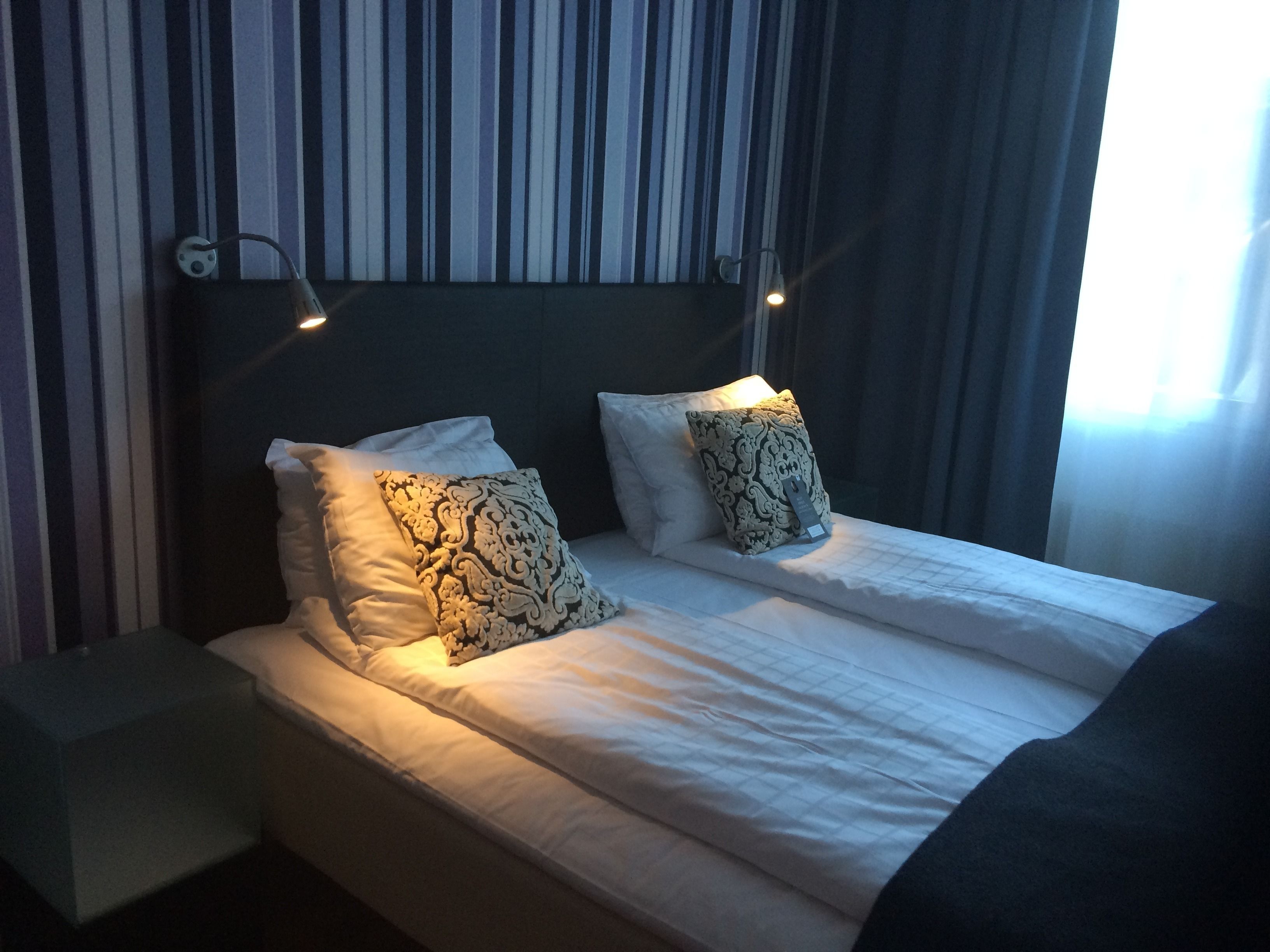 A neatly made bed with white linens and two patterned pillows, a bedside table, and a wall with striped wallpaper.