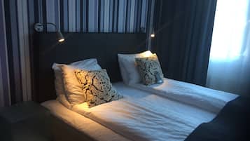 Una cama bien hecha con sábanas blancas y dos almohadas estampadas, una mesita de noche y una pared con papel tapiz a rayas.