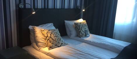 A neatly made bed with white linens and two patterned pillows, a bedside table, and a wall with striped wallpaper.