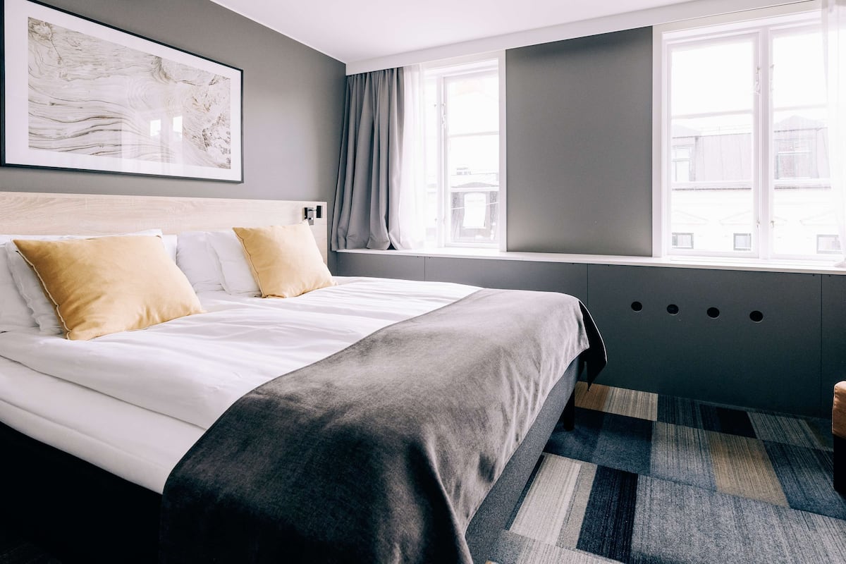 A modern hotel room with a large bed, a window with curtains, a headboard, and a wall-mounted artwork.