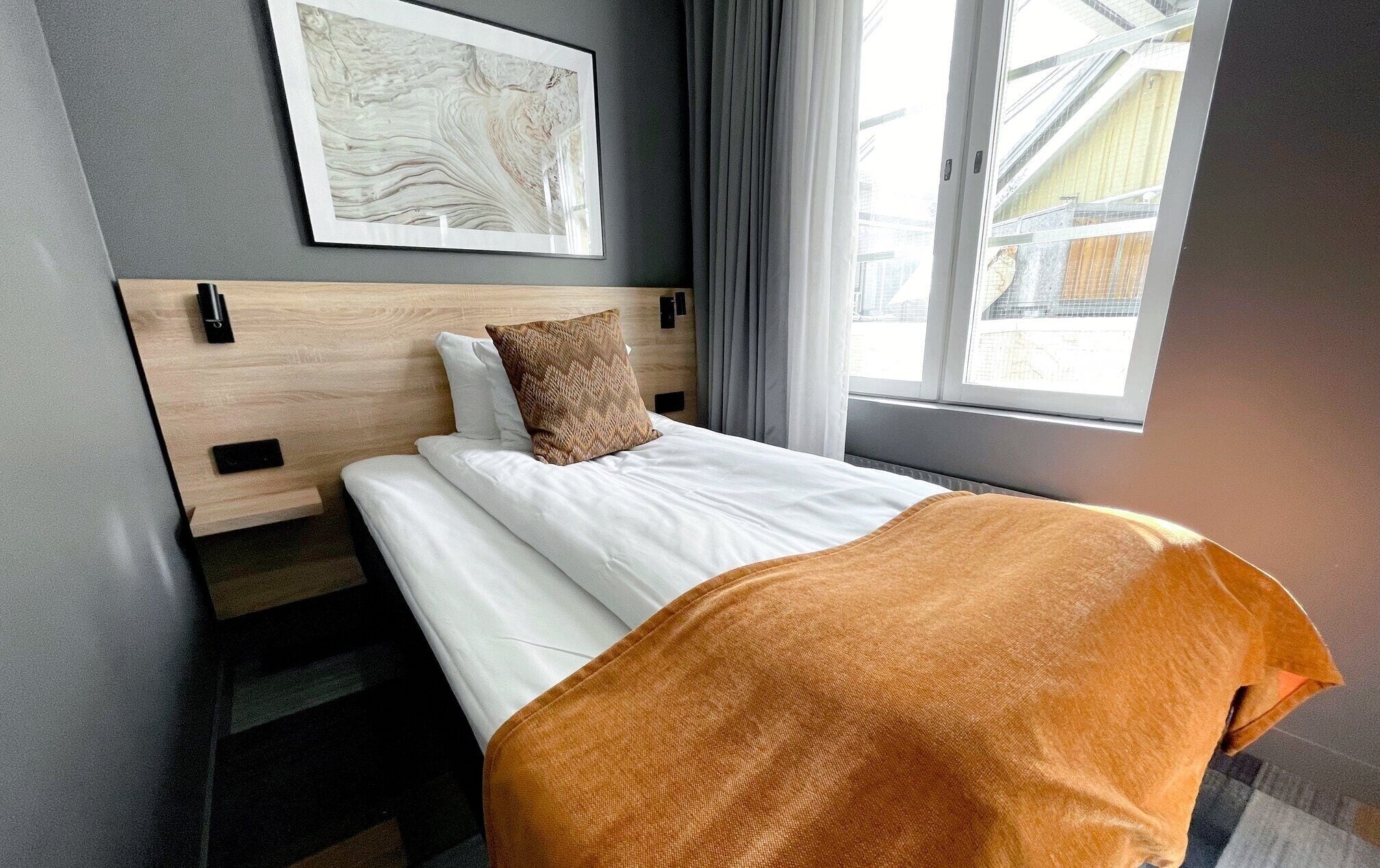 Ein Hotelzimmer mit Bett, Kopfteil, Fenster und einem an der Wand hängenden Kunstwerk.