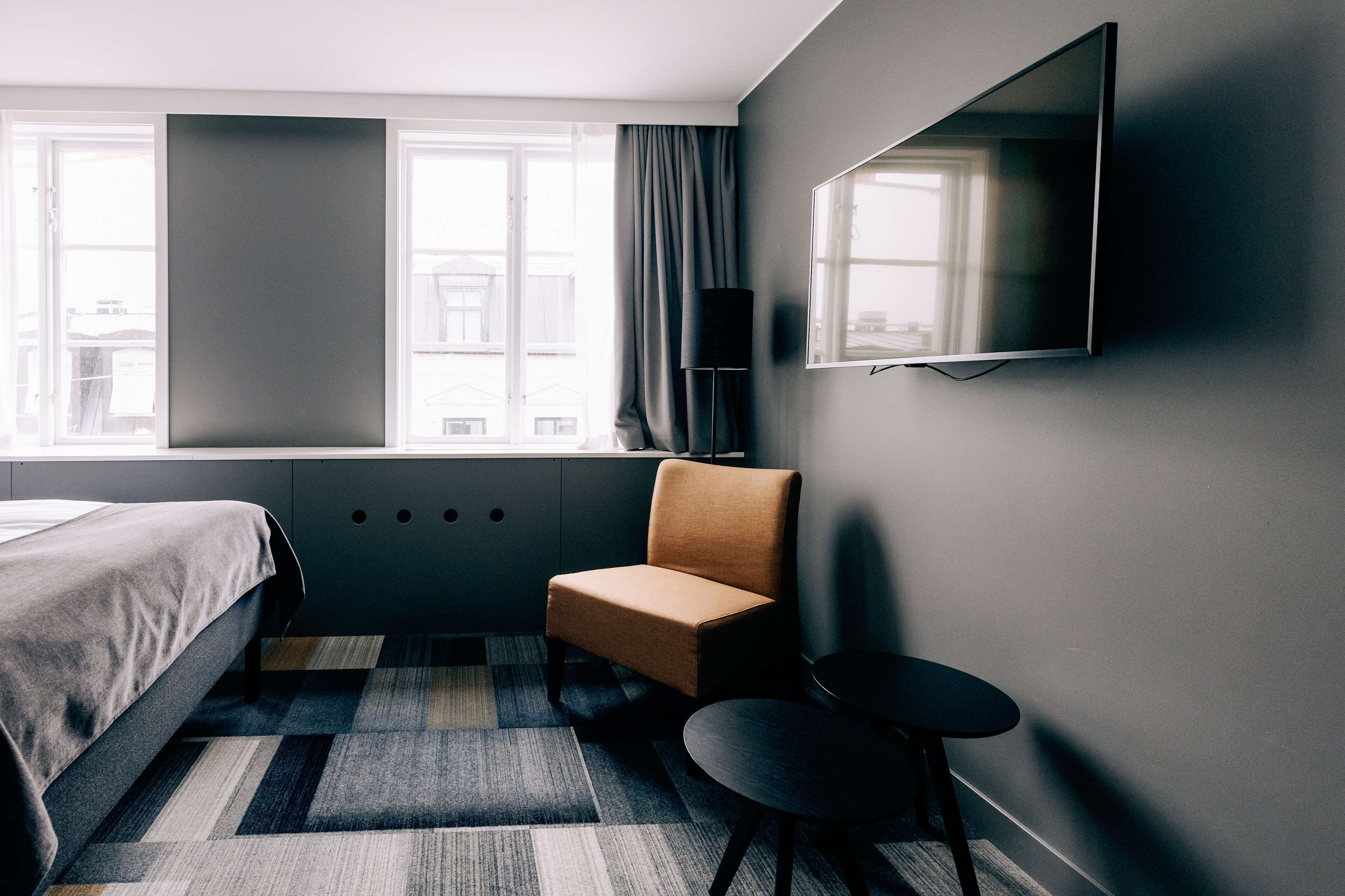 Ein Hotelzimmer mit Bett, Sessel, kleinem Tisch, Wandfernseher und einem Fenster mit Vorhängen.