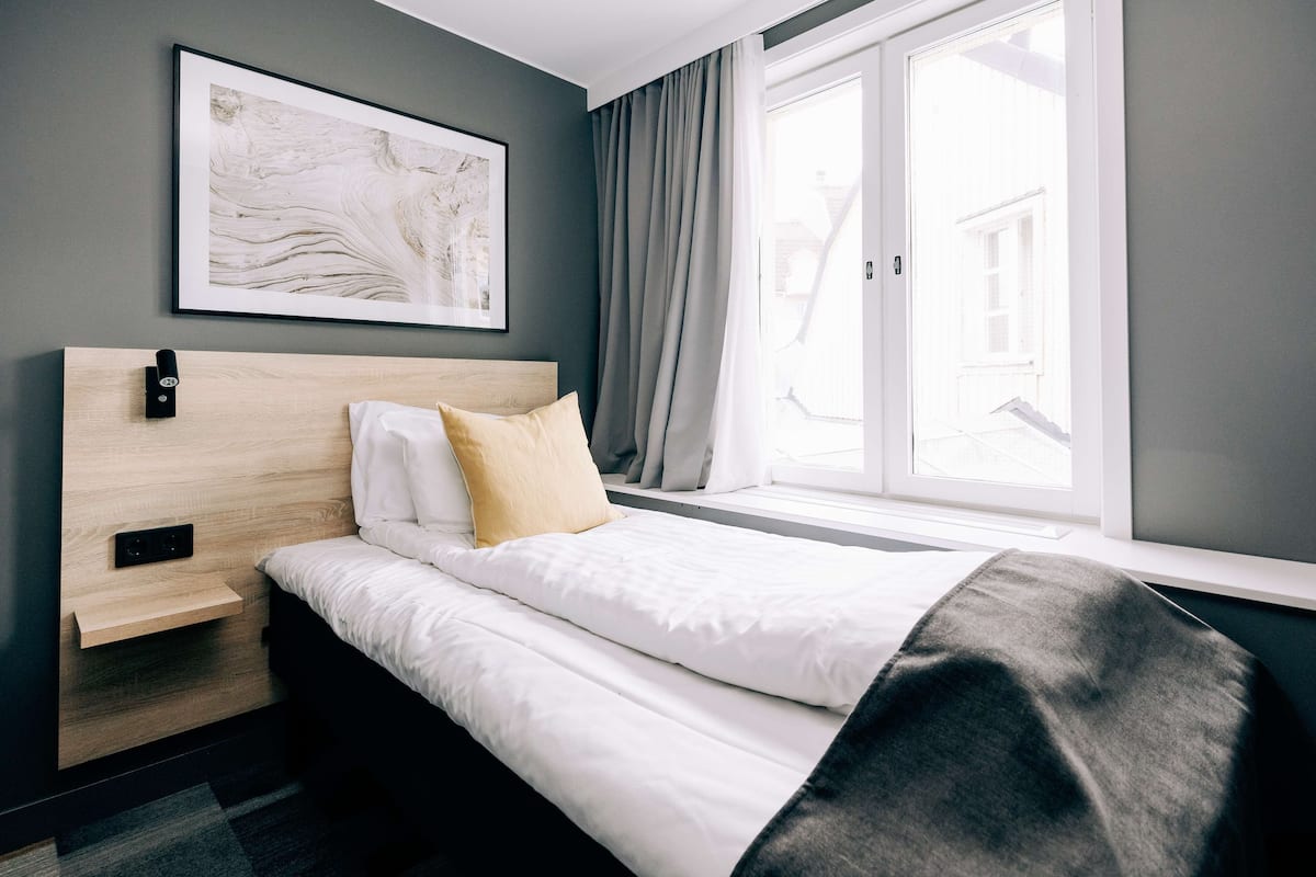 A modern hotel room with a bed, a bedside table, a framed artwork, and a window with curtains.