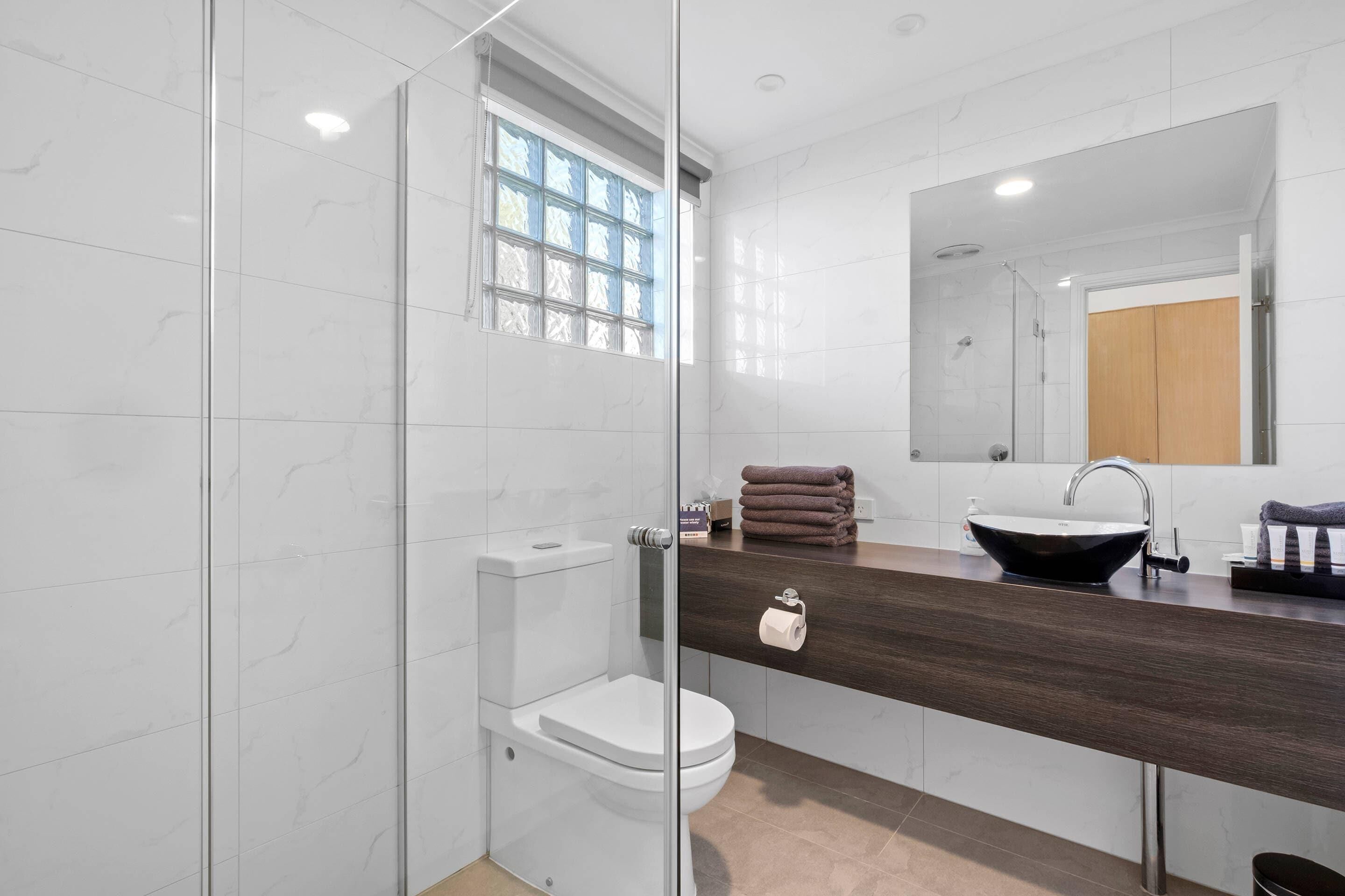A modern bathroom with a glass-enclosed shower, a white toilet, a dark wood vanity, a large mirror, and a black sink.