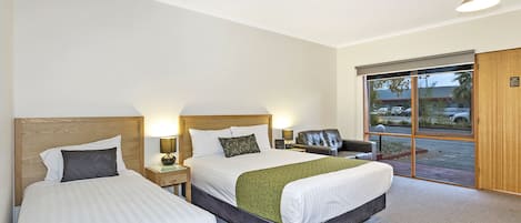 A hotel room with two beds, a wooden headboard, a green throw blanket, and a view of the outside through a large window.