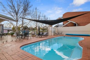 A swimming pool with a brick deck, surrounded by outdoor furniture and a shaded area.