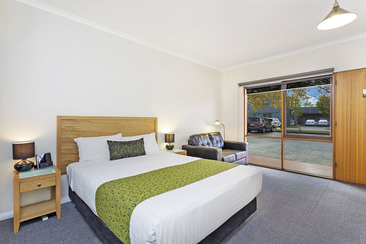 A hotel room with a large bed, a wooden headboard, a green patterned bedspread, a leather armchair, a nightstand with a lamp, and a sliding glass door leading to a parking area.