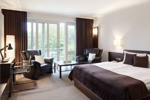 A hotel room with a large bed, two armchairs, a small table with fruit, a desk with a lamp, and a view of trees through the windows. - Nordica Hotel Berlin (Heide)