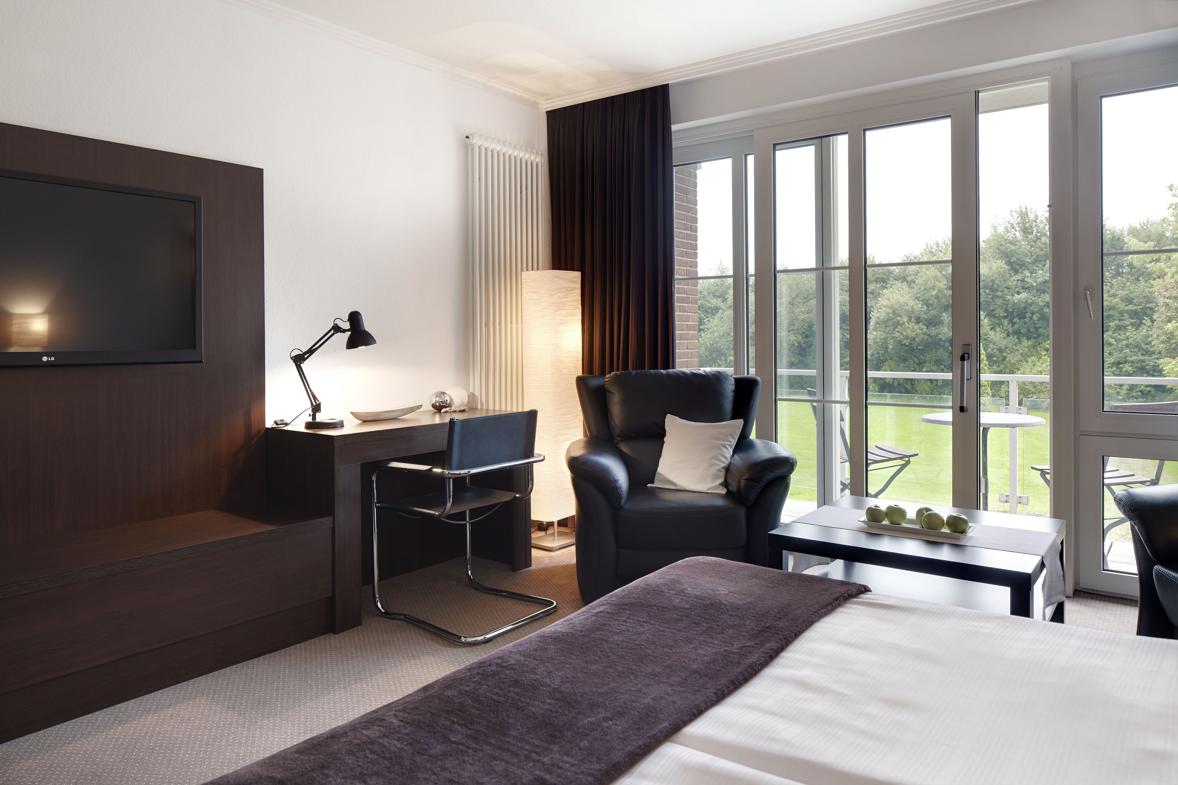 Quarto de hotel moderno com cama, escrivaninha, cadeira, televisão e janelas amplas que oferecem vista para o exterior.