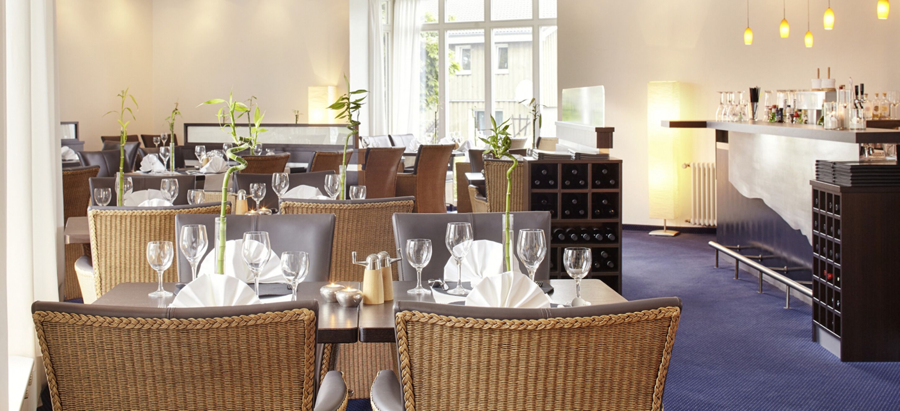 En restaurant med fletstole, borde dækket med vinglas og tallerkener, samt en bar med vinflasker.