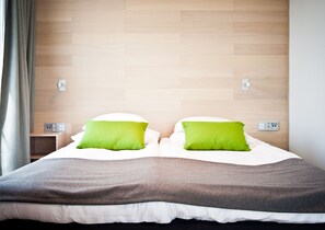 Een moderne hotelkamer met een houten hoofdbord, twee groene kussens, wit beddengoed en een nachtkastje met een lamp.
