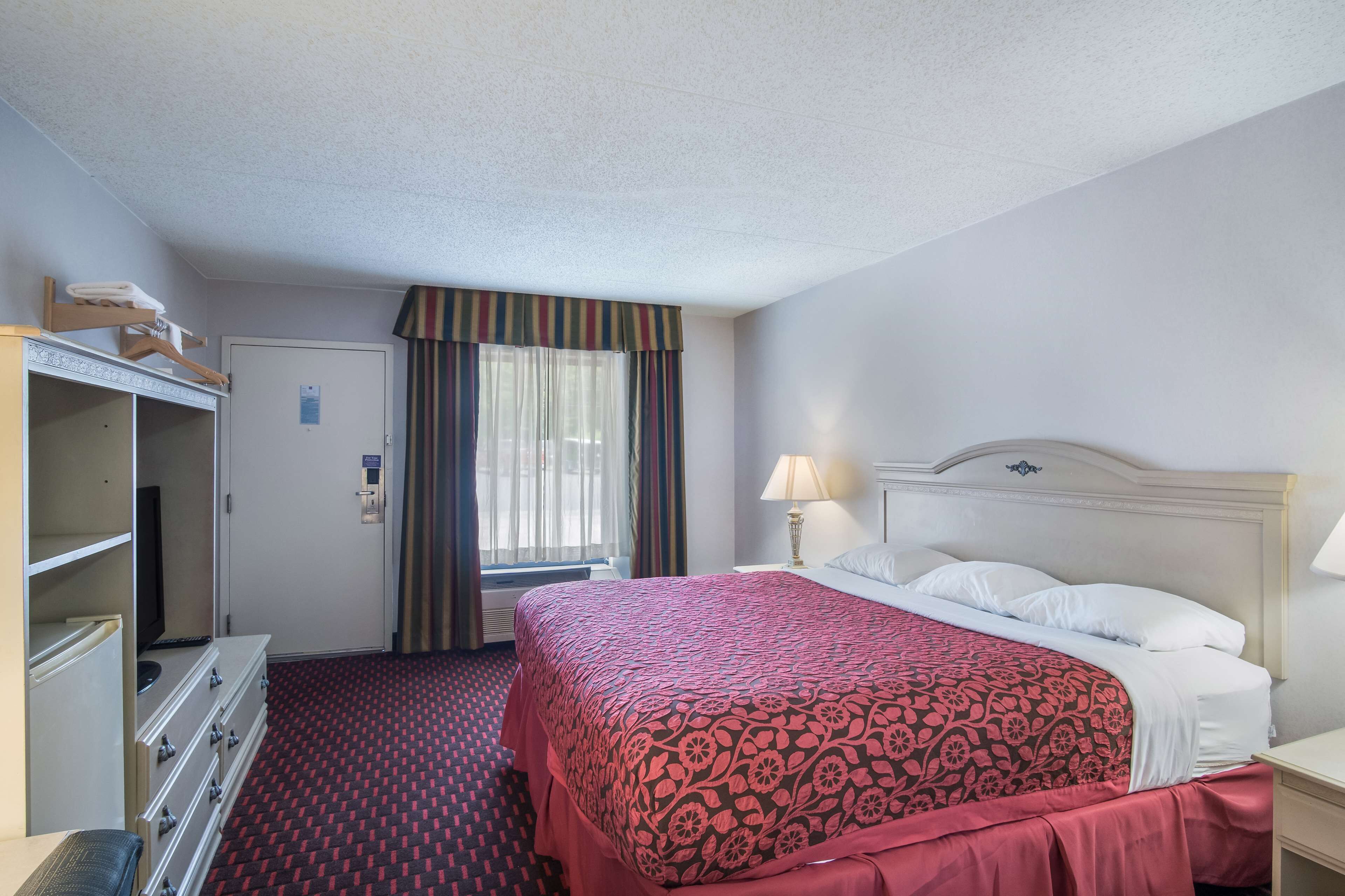 Une chambre d’hôtel comprenant un lit, une commode, un téléviseur, une fenêtre avec des rideaux et une lampe.