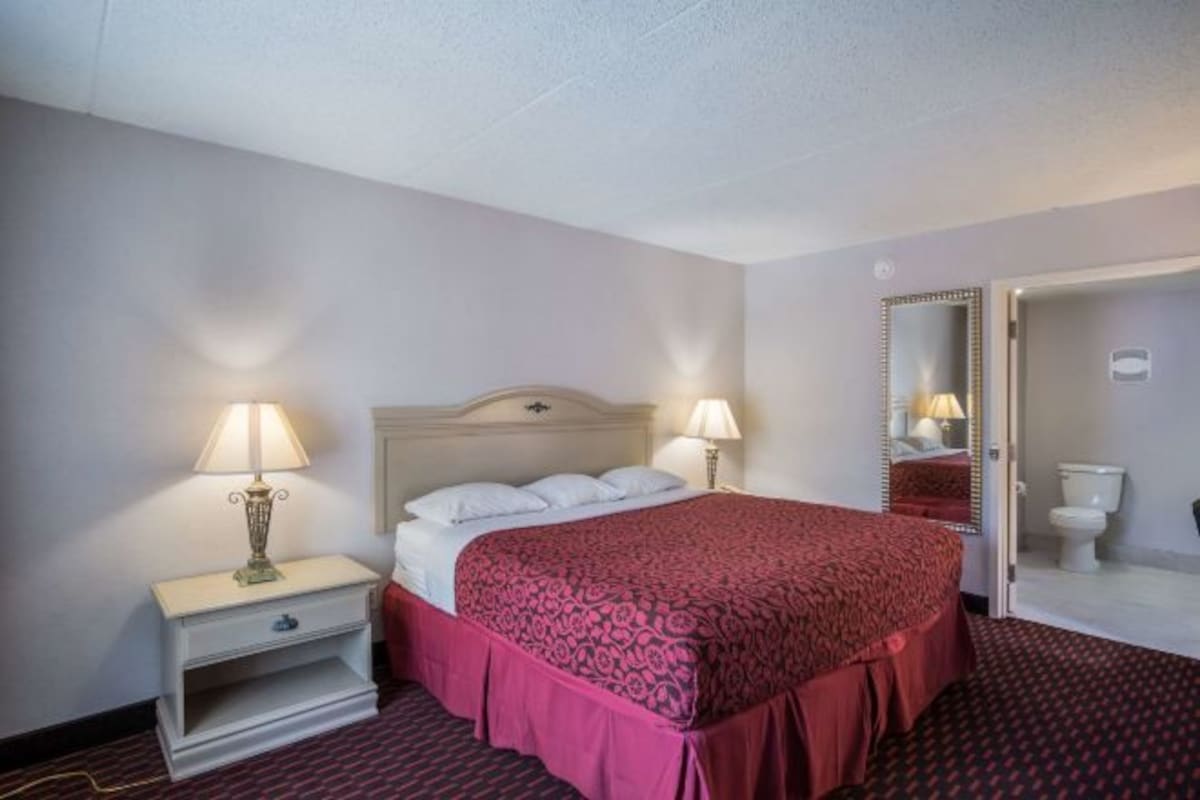 A hotel room with a bed, two bedside lamps, a nightstand, and a bathroom visible through an open door.