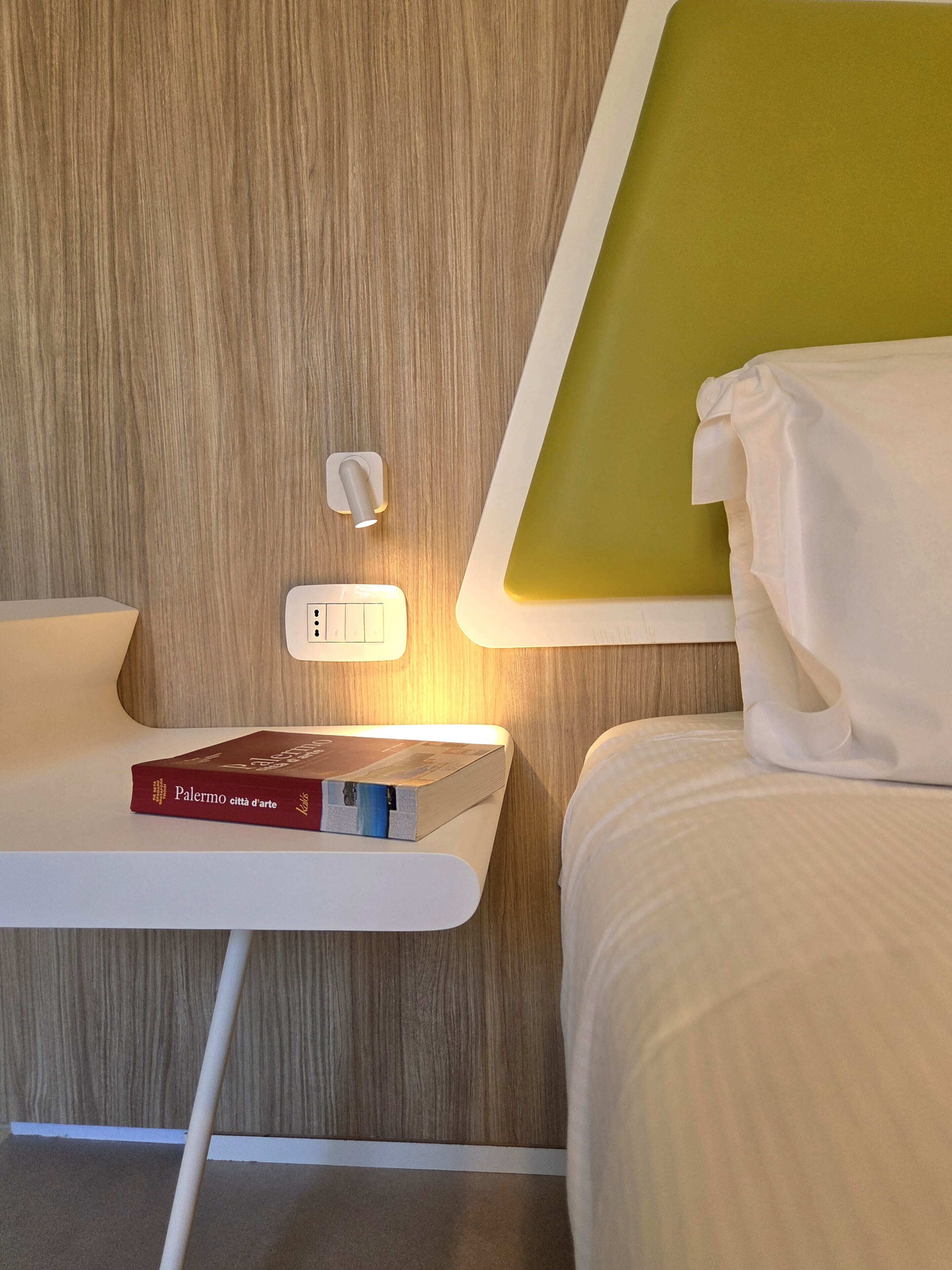 A neatly arranged hotel room with a bed, a bedside table with a book, a wall-mounted light, and a power outlet.