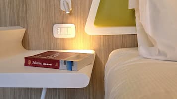 A neatly arranged hotel room with a bed, a bedside table with a book, a wall-mounted light, and a power outlet.