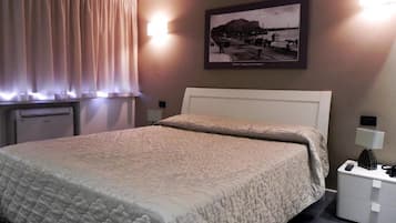 A hotel room with a bed, a nightstand, a lamp, a framed picture on the wall, and a window with curtains.