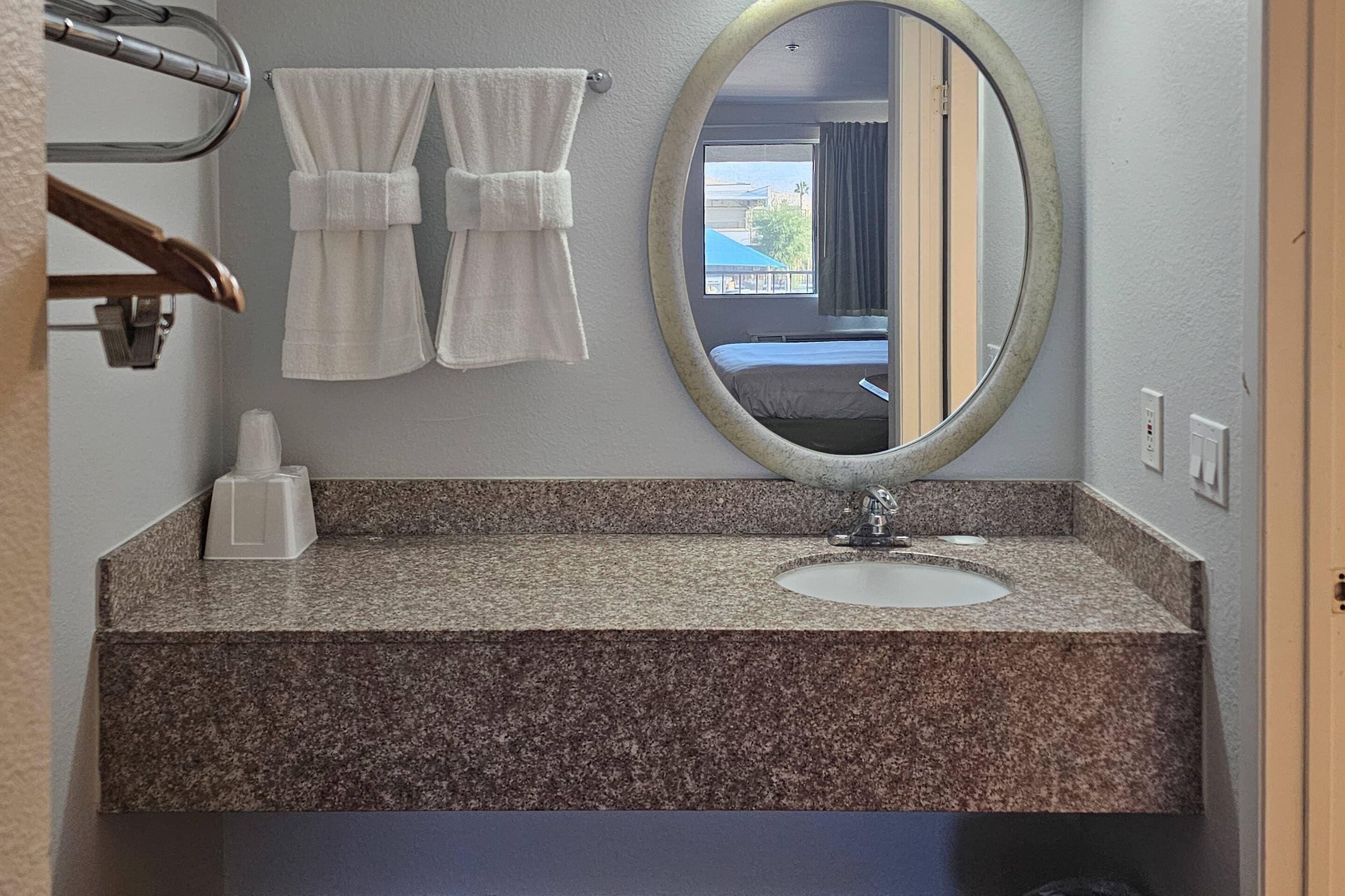 A bathroom with a granite countertop, a white sink, a round mirror, and two towels hanging on a towel rack.