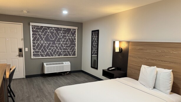 Une chambre d’hôtel comprenant un lit, un bureau, une chaise, un radiateur, un téléphone fixé au mur et une fenêtre ornée d’un motif géométrique.