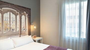 Habitación de hotel con una cama grande, una mesita de noche, una lámpara y vistas a edificios a través de la ventana.