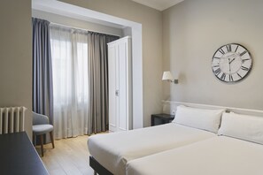 A hotel room with a bed, a nightstand, a lamp, a chair, and a view of the outside through a window with curtains.