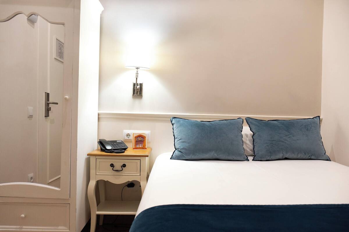 Ett hotellrum med en säng, ett nattduksbord med en telefon och en lampa samt en dörr.