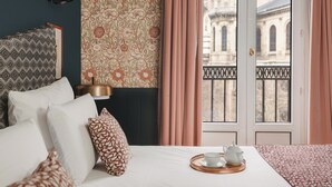 Ein Hotelzimmer mit Bett, Kissen, Nachttisch mit Lampe, Tablett mit Teekanne und Tasse sowie Blick auf ein Gebäude durch ein mit Vorhängen versehenes Fenster.
