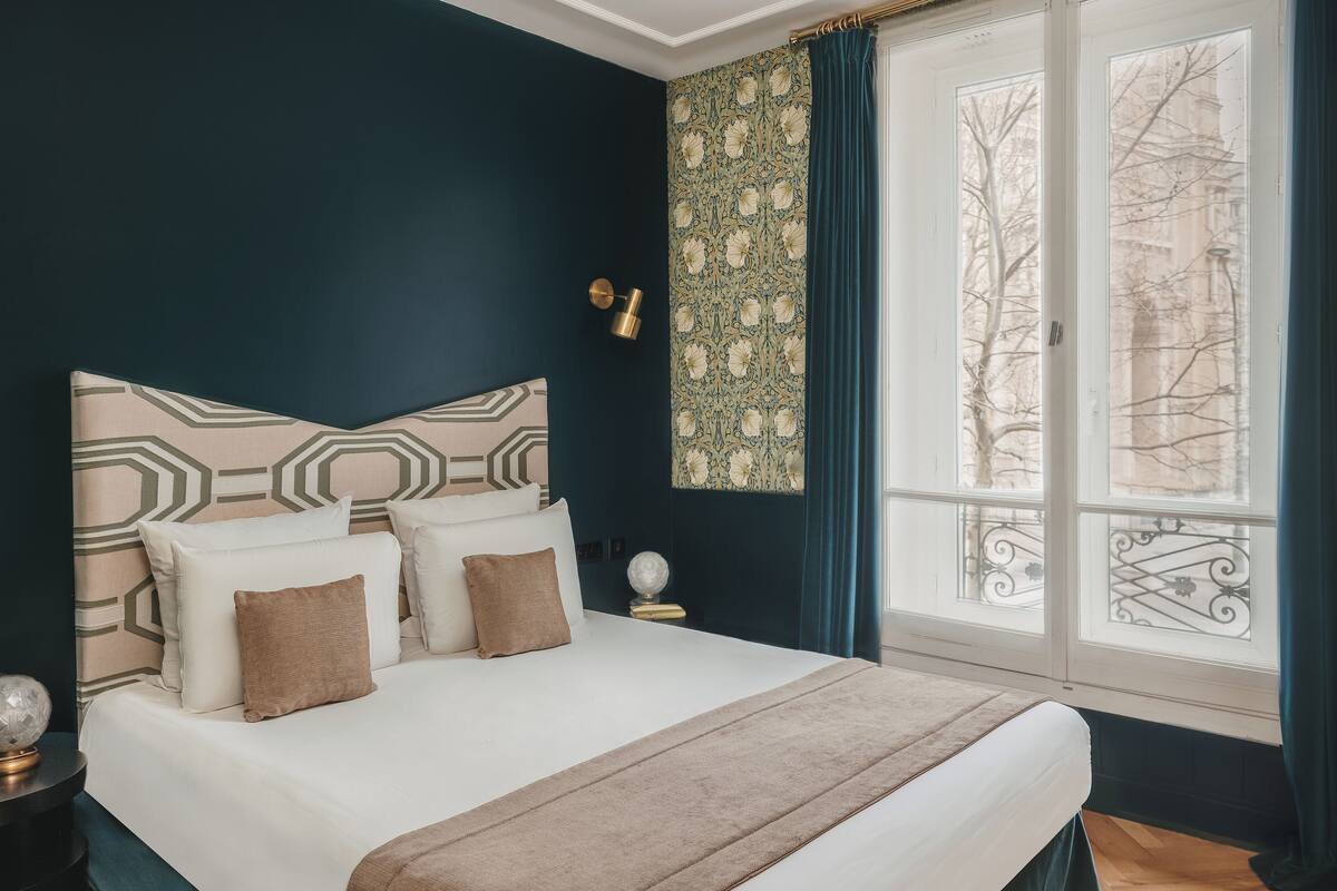 A neatly made bed with a geometric-patterned headboard, a bedside table with a lamp, and a view of a building through a window with a curtain.