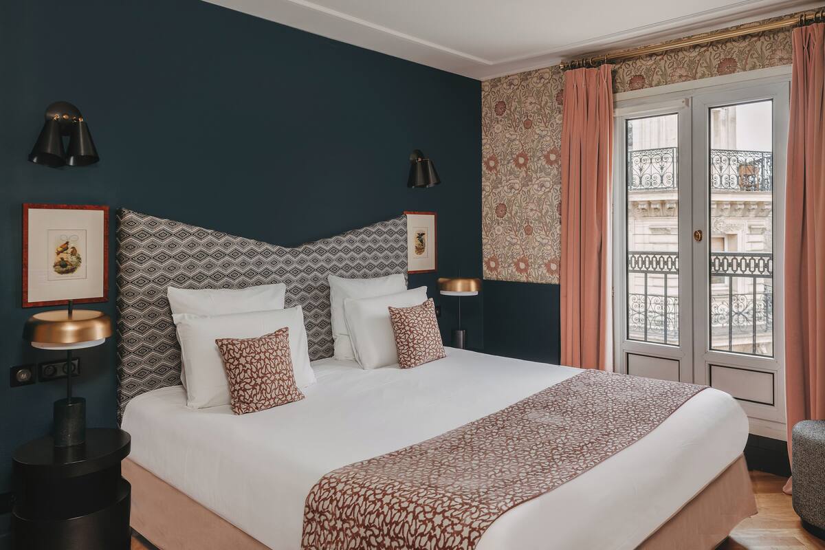 A neatly arranged bedroom with a large bed, a bedside table, a lamp, and a view of the outside balcony.