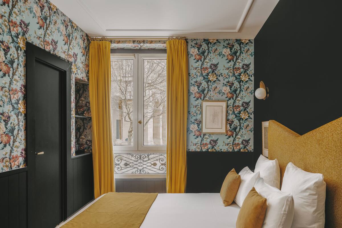 A bedroom with floral wallpaper, a bed with white and mustard pillows, and a window with yellow curtains.