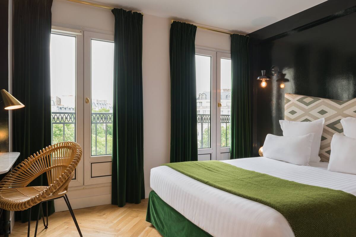 A hotel room with a bed, a chair, a desk, and a view of the city through large windows.