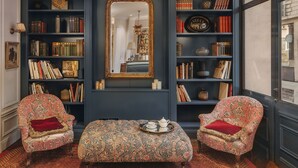 Ein Zimmer mit zwei gemusterten Sesseln, einem passenden Hocker, einem Bücherregal voller Bücher, einem großen Spiegel und einer dekorativen Lampe.