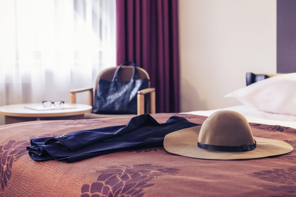 Ein Hotelzimmer mit einem Bett, einem runden Tisch mit einem Stuhl, einem Koffer und einem Hut auf dem Bett.