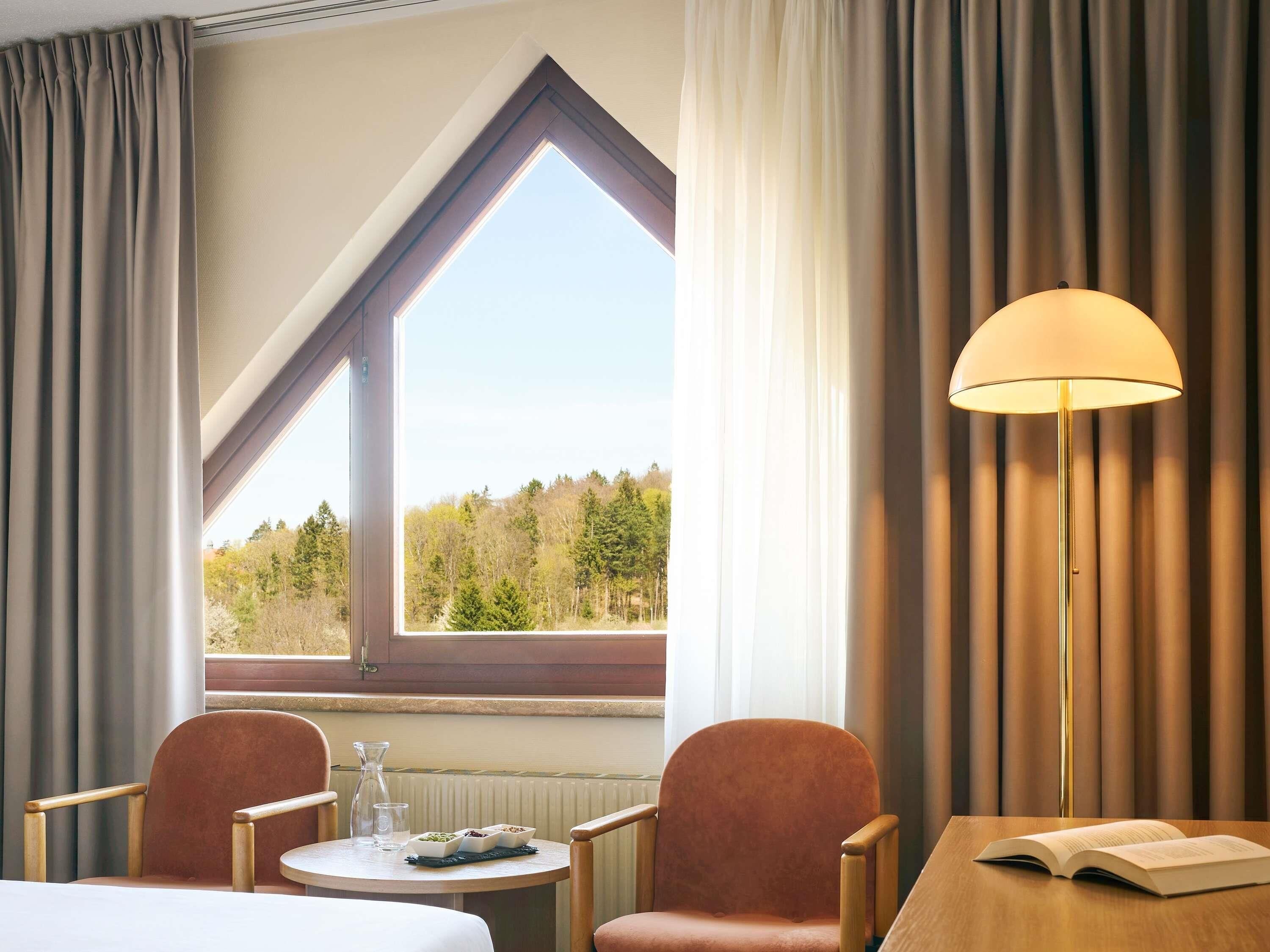Ein Hotelzimmer mit großem Fenster, zwei Sesseln, einem kleinen Tisch mit Buch und Glas sowie einer Stehlampe.