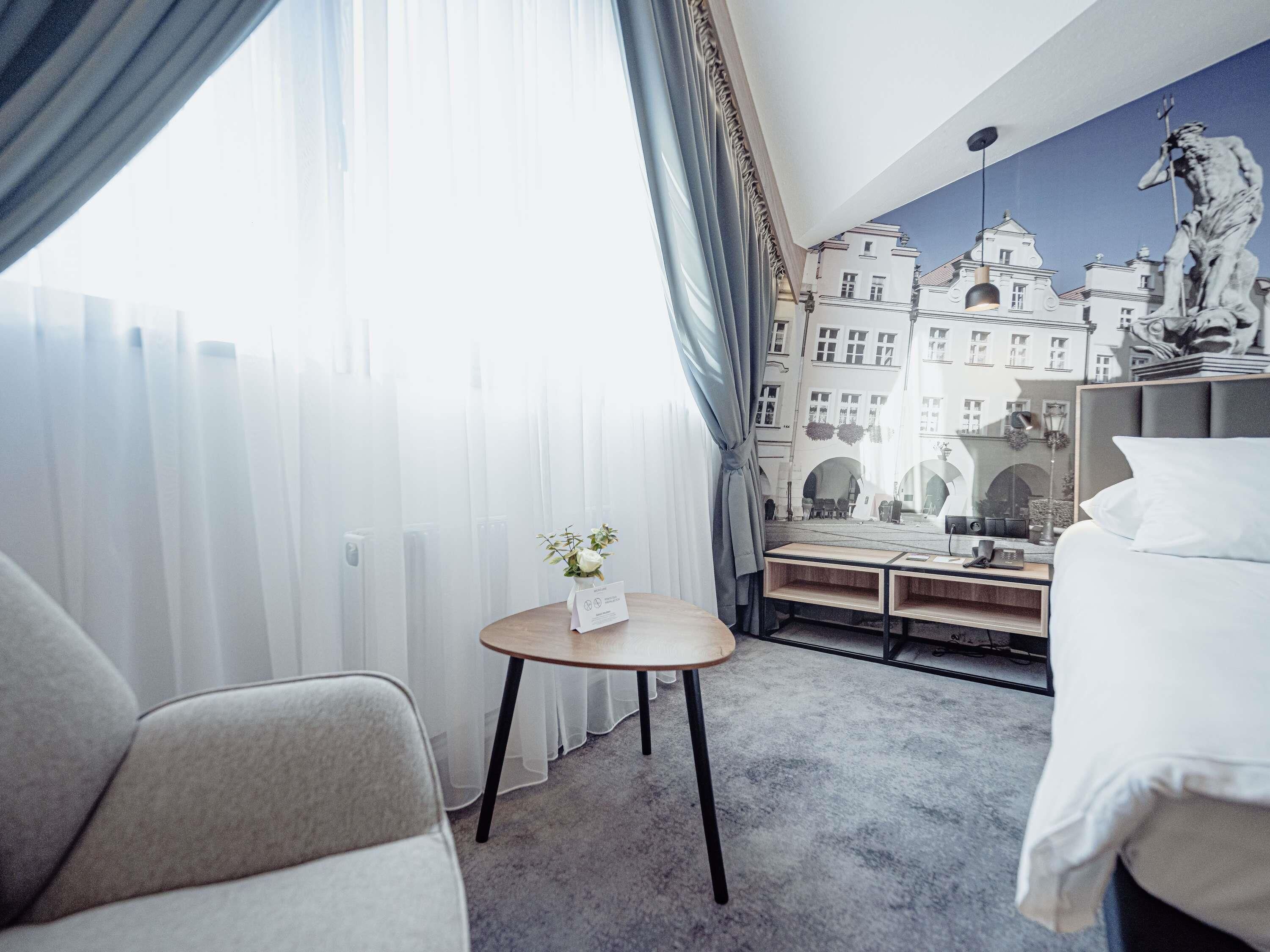 A hotel room with a bed, a chair, a small table with a vase, and a view of buildings through a window.