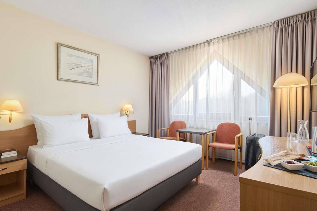 Ein Hotelzimmer mit Bett, Nachttischen, Schreibtisch, Stuhl und einem Vorhangfenster.