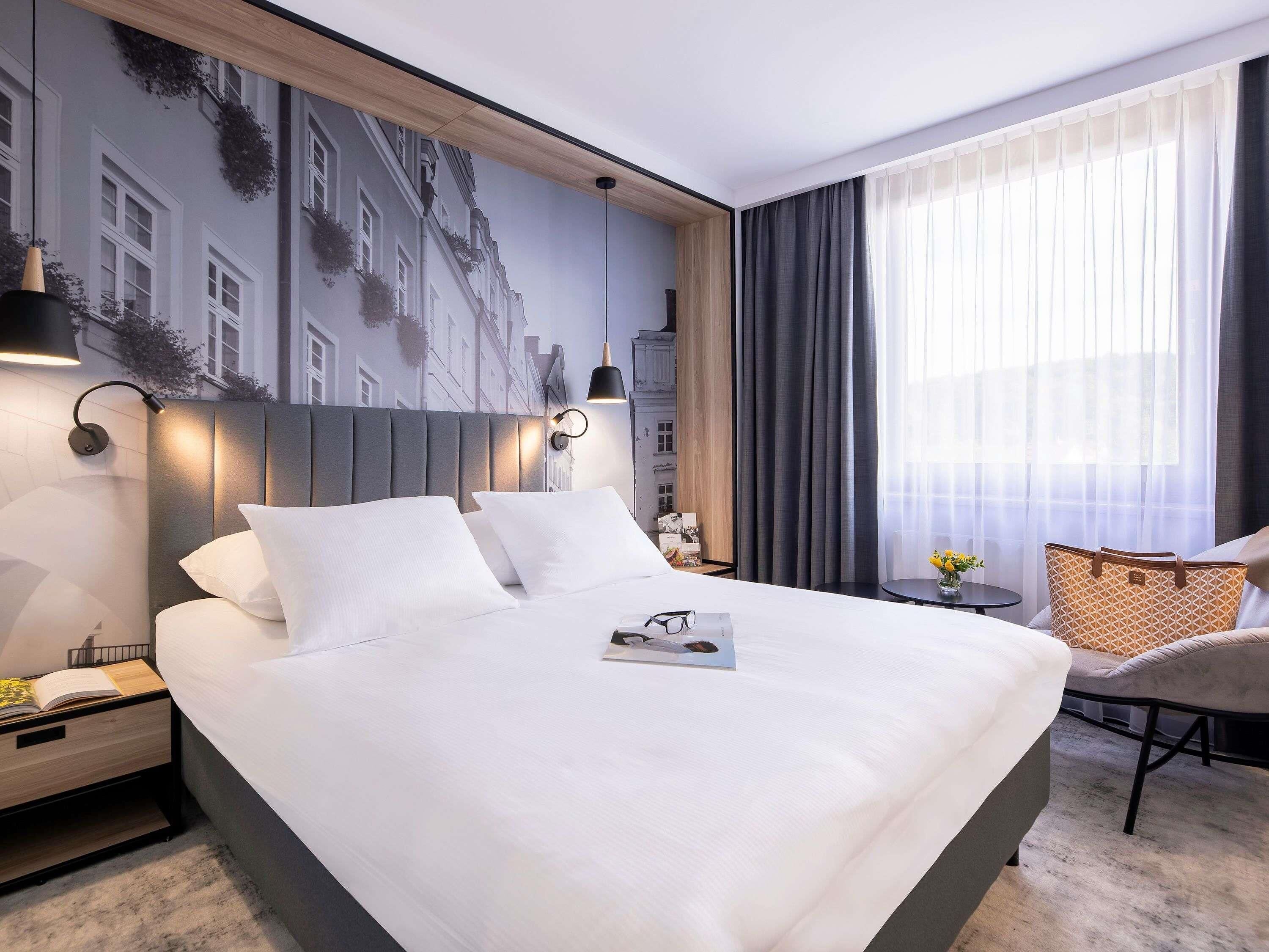 Una habitación de hotel moderna con una cama grande, una mesita de noche, una lámpara de lectura, una lámpara de pared, una silla y una ventana con cortinas.