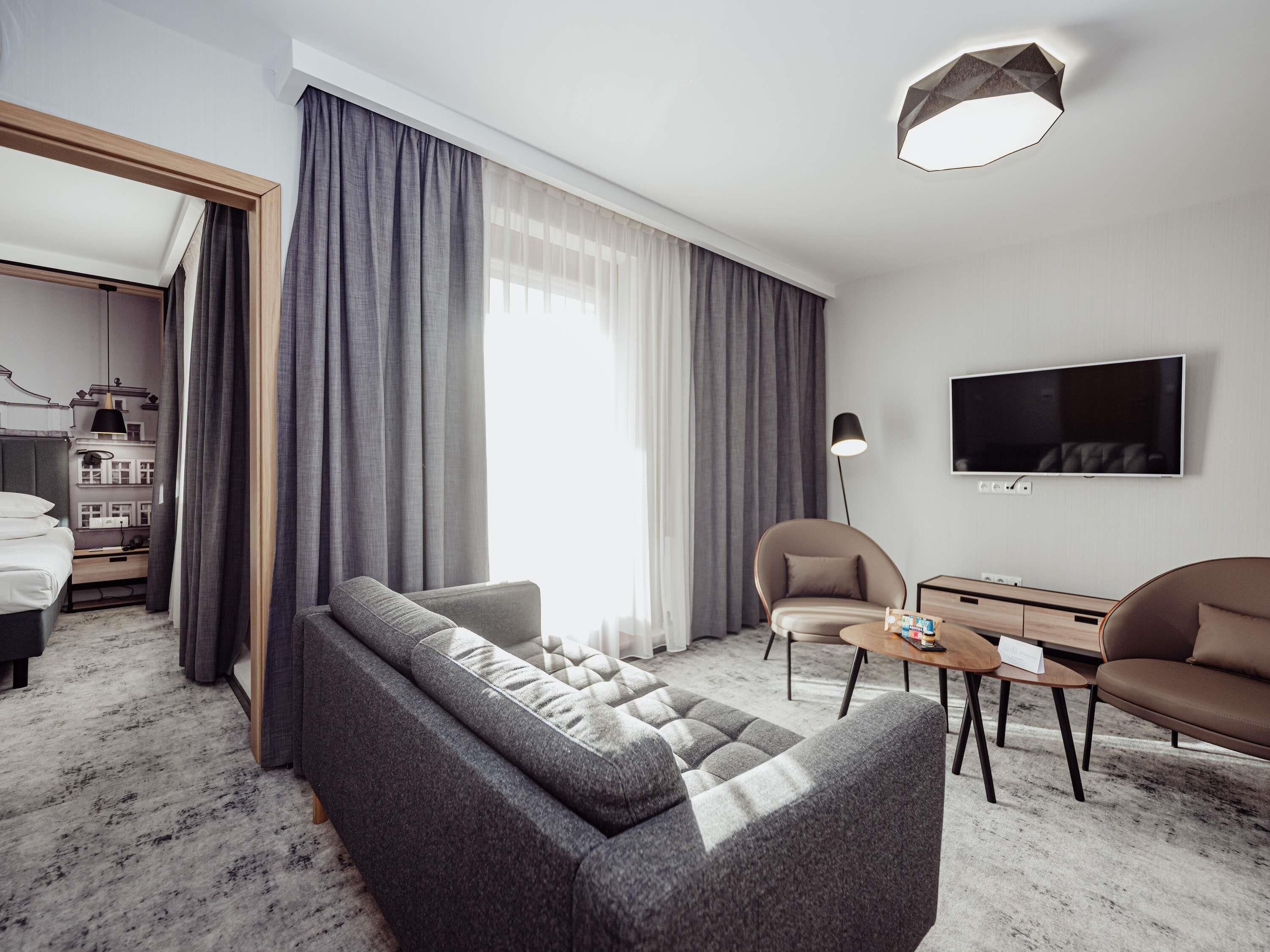 A modern hotel room with a grey sofa, a small round table, a TV, and a large window with curtains.