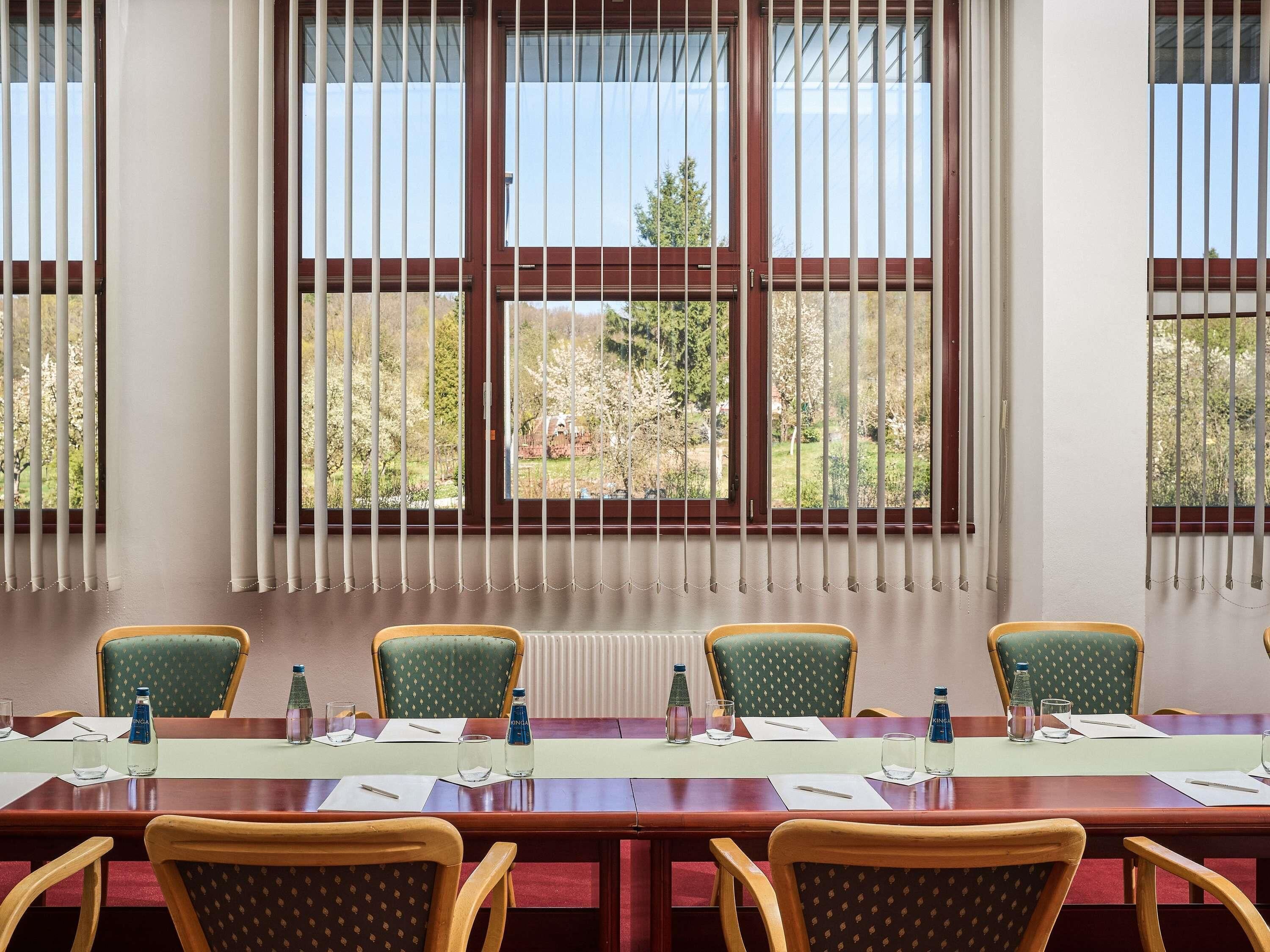 A conference room with a long table, chairs, and a view of trees through barred windows.