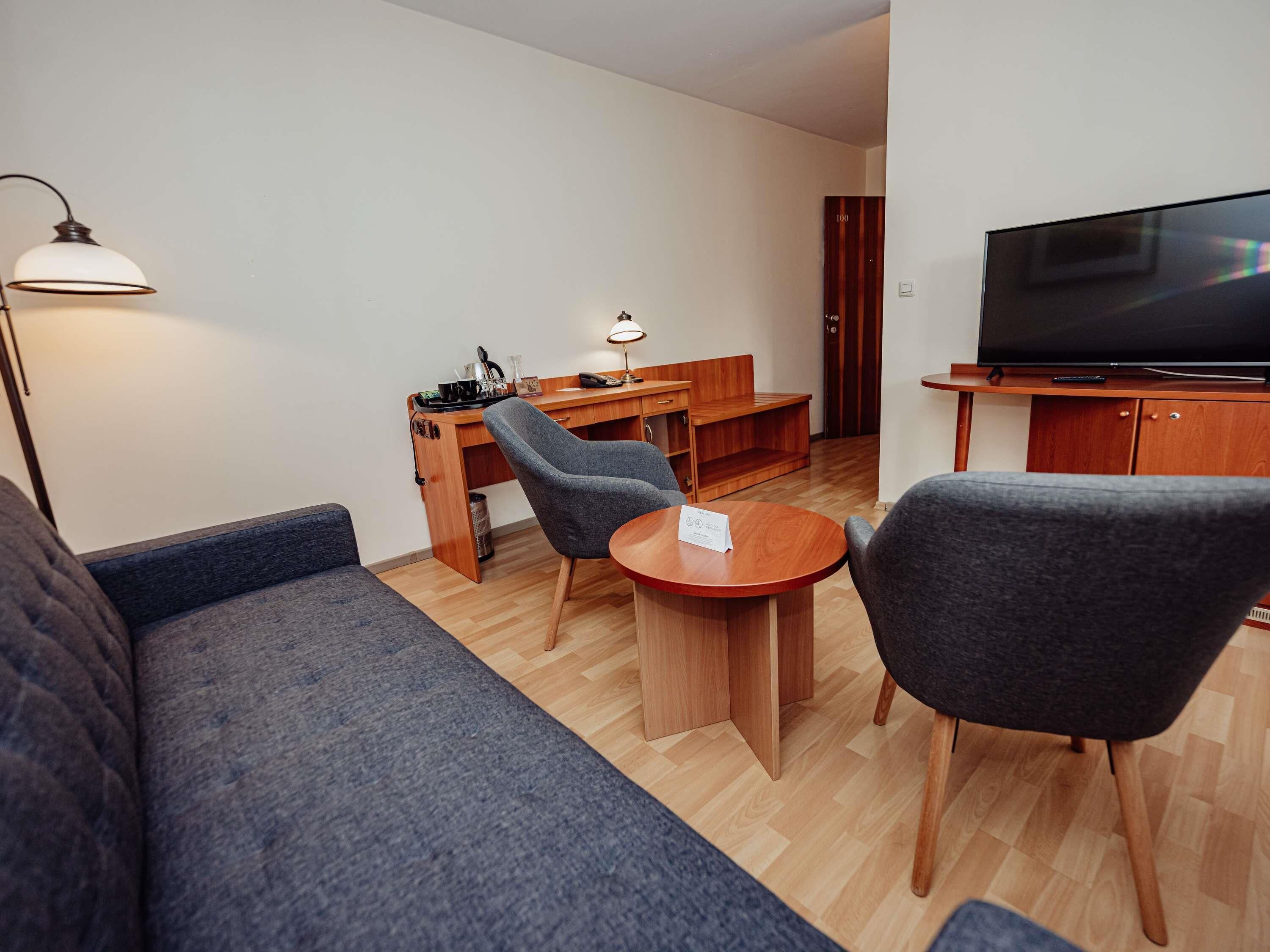A modern living room with a sofa, two armchairs, a coffee table, a desk with a lamp, a television, and a door.