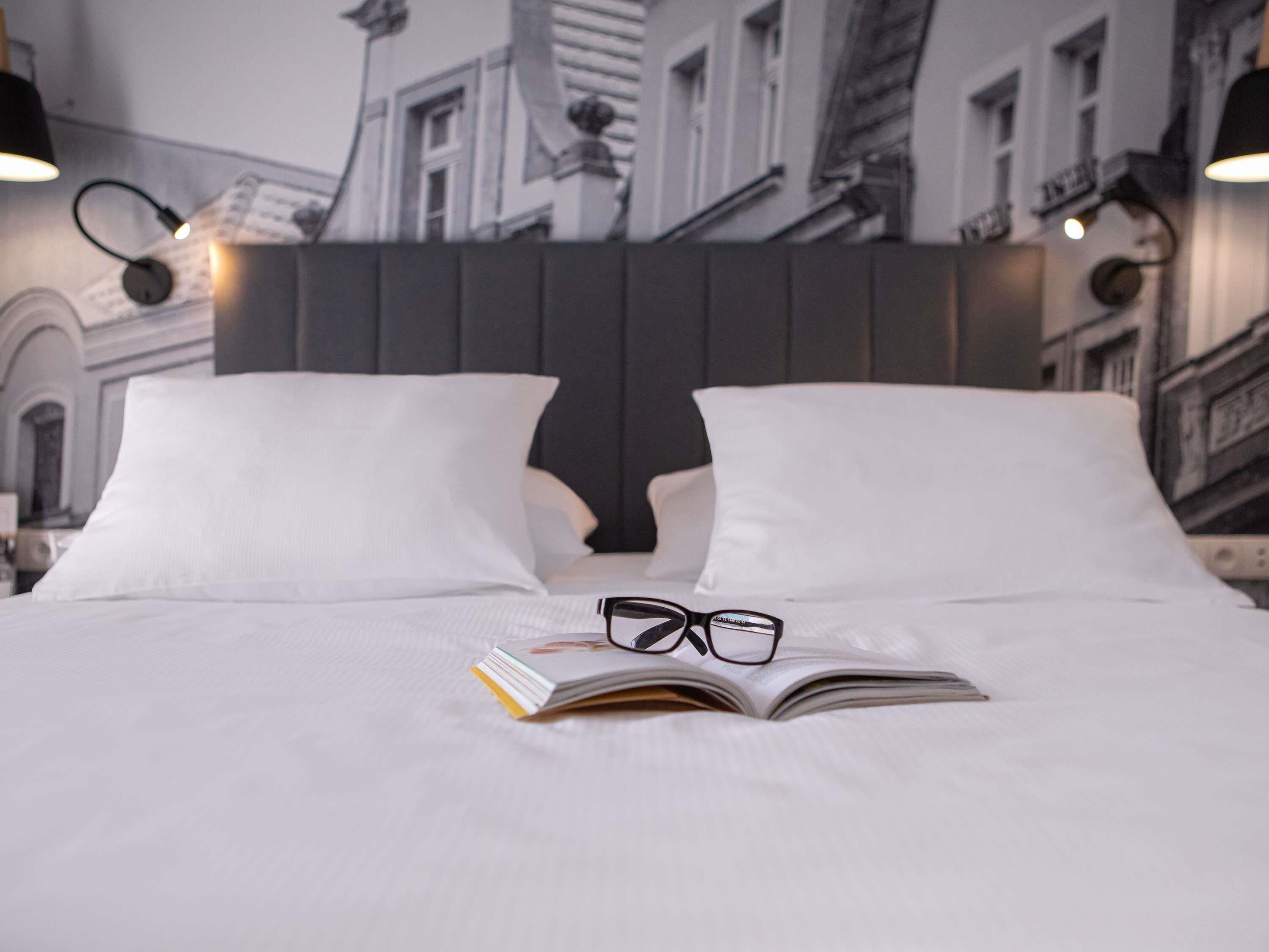 A neatly made bed with white linens, a book, and glasses on it, set against a backdrop of a cityscape wallpaper.