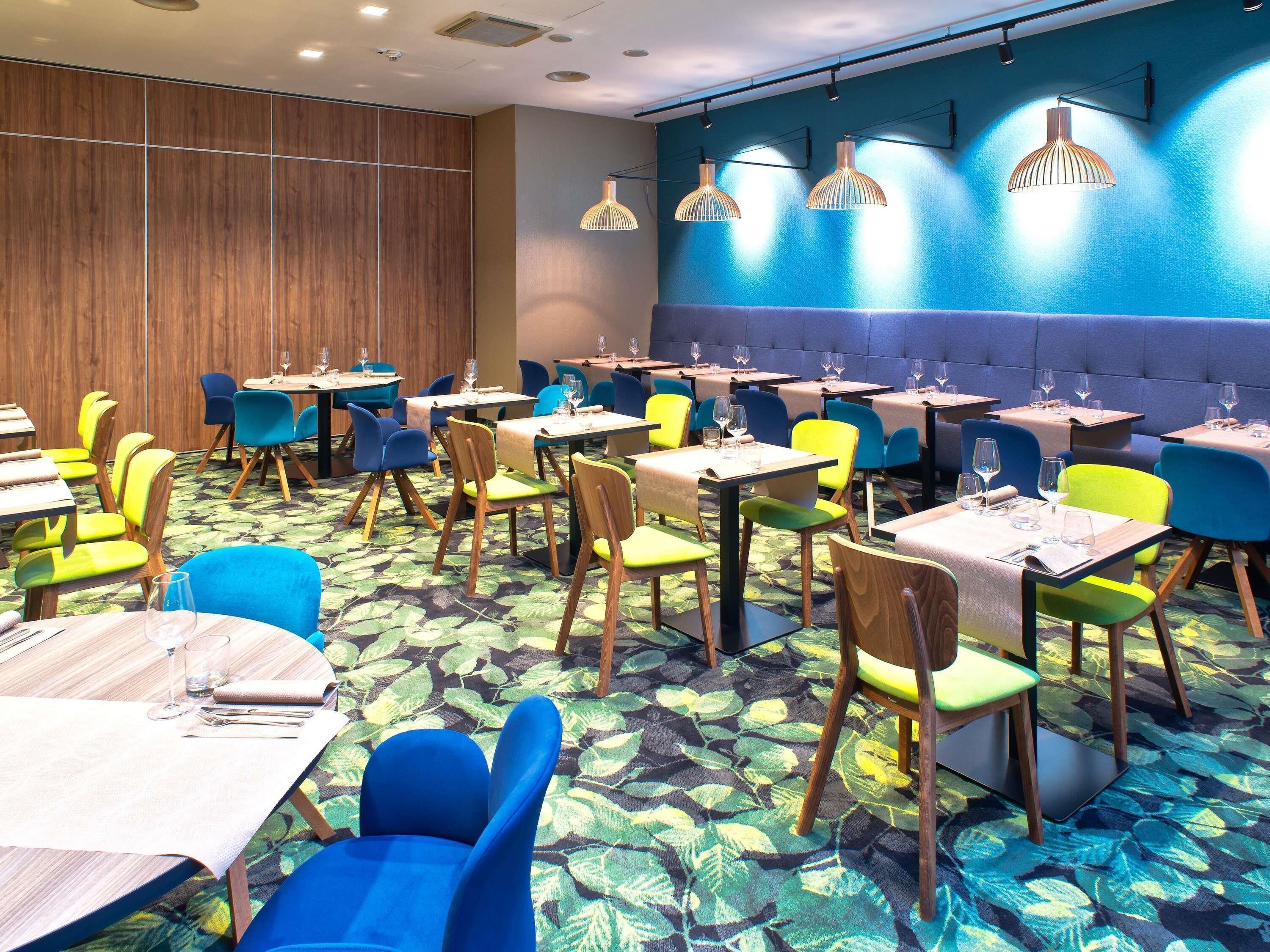 Un comedor con mesas y sillas, alfombra estampada y pared azul con lámparas colgantes.
