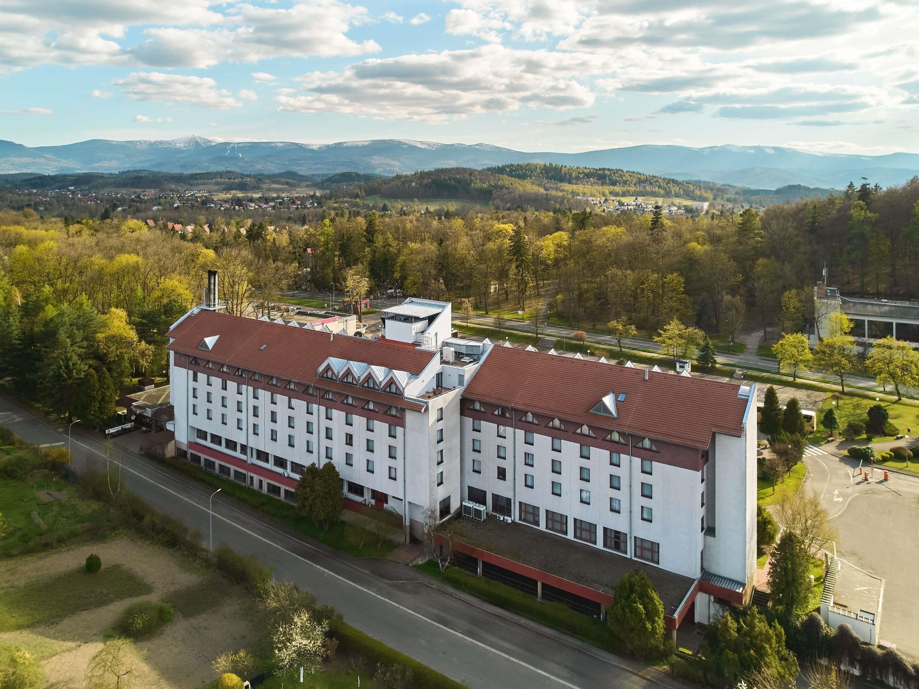 A large, multi-story hotel with a red-tiled roof, surrounded by a lush green landscape and mountains in the background.
