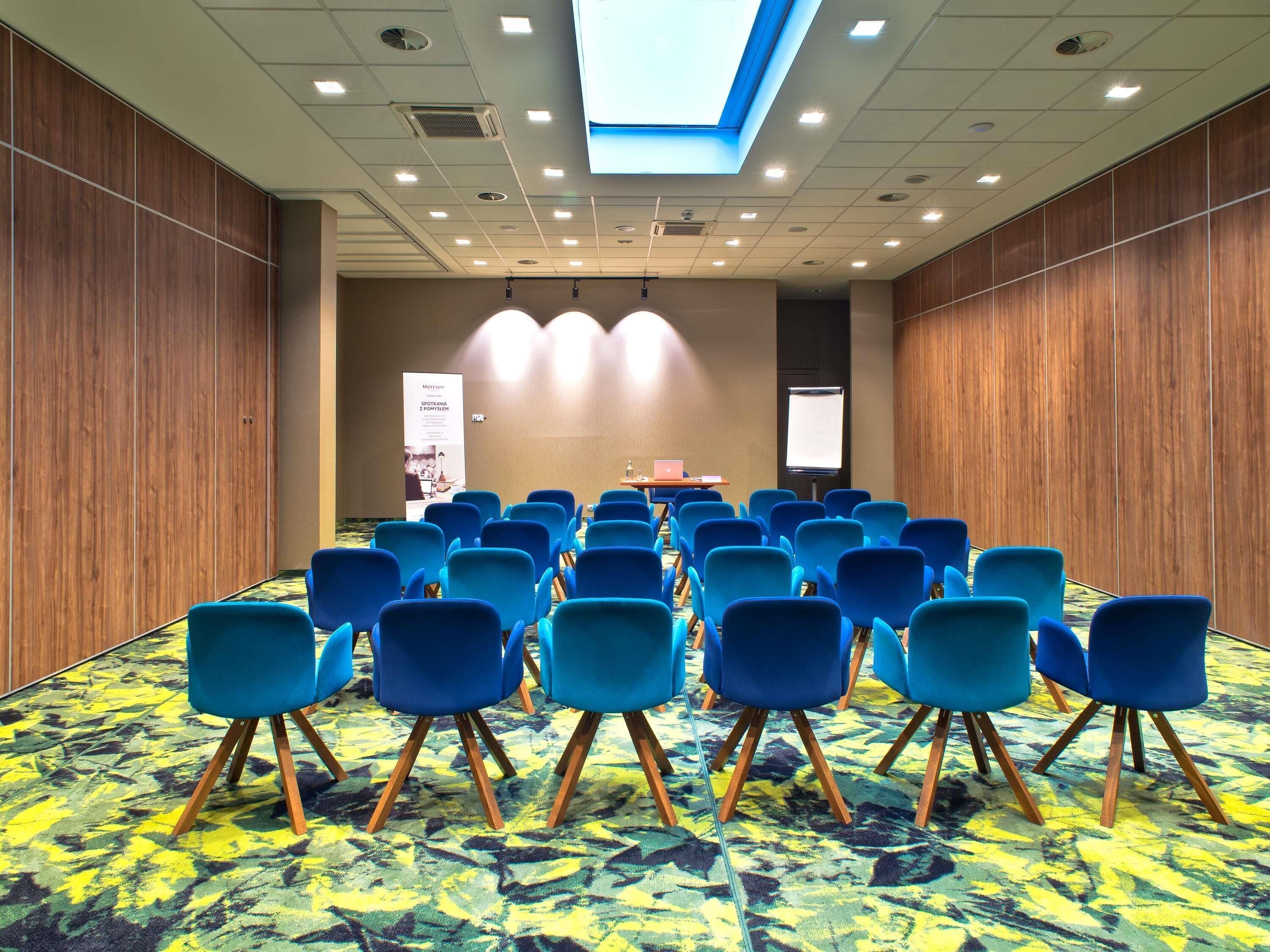 Un salón de conferencias con hileras de sillas azules, un atril, una pantalla y un cartel.